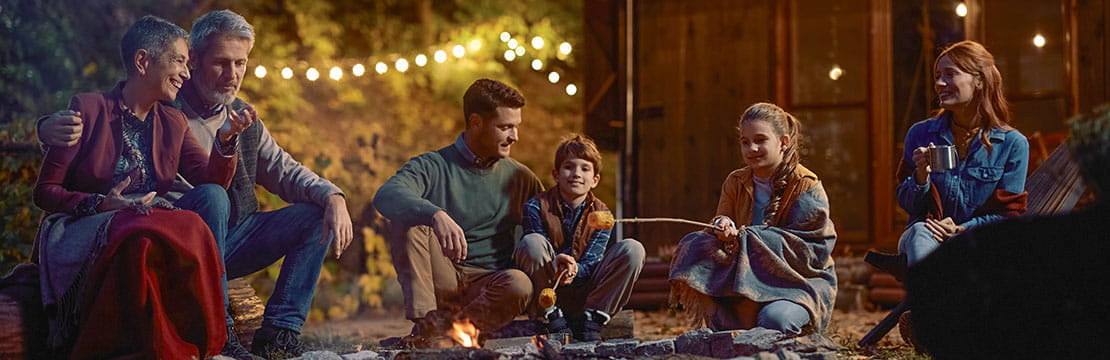 Happy multigeneration family gathering around campfire roasting corn in the evening.